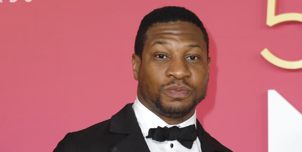 jonathan majors, a man stands looking at the camera holding his hands together, close cropped black hair, wearing a black suit and bow tie with white shirt