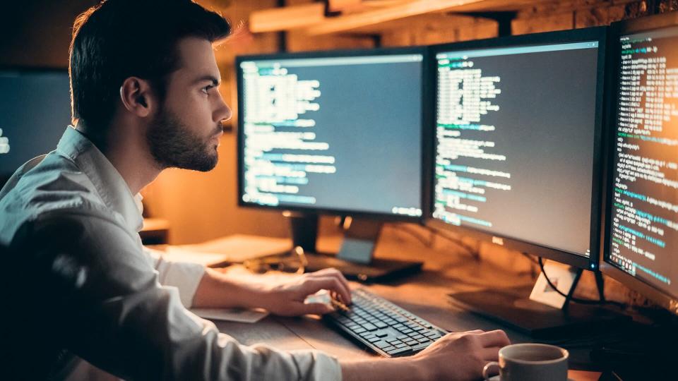 Focused young developer coding late in office writing script shown on computer monitors, serious handsome coder programmer hacker programming developing software applications working alone at night.