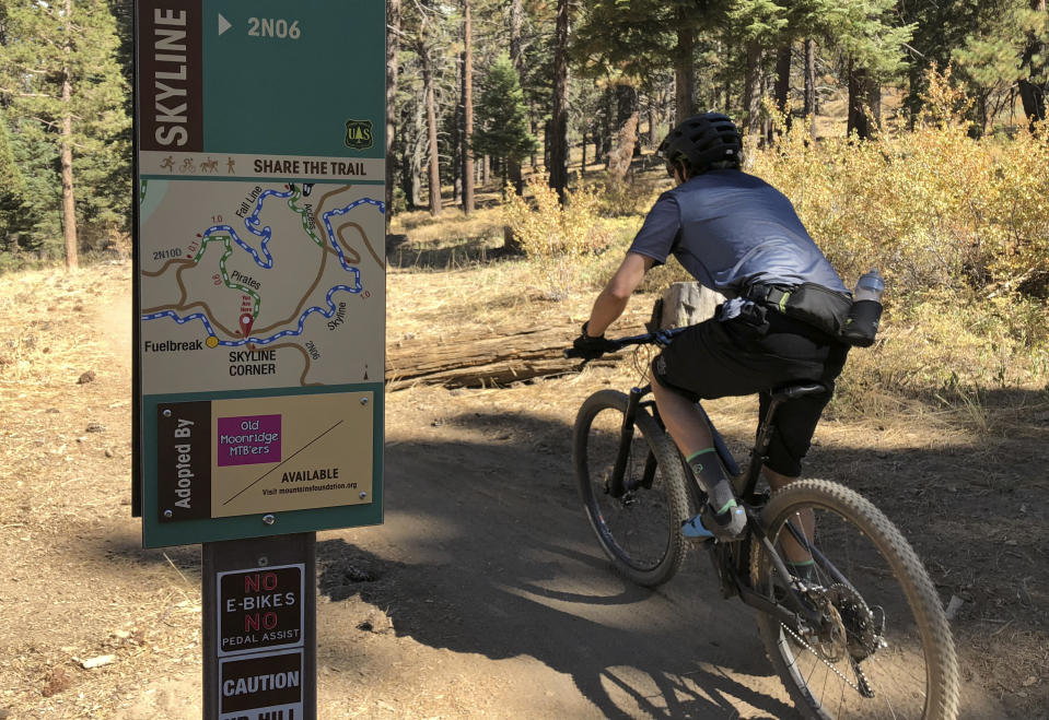 In this photo taken Sept. 23, 2018, a mountain biker pedals past a No E-bikes sign in the San Bernardino National Forest near Big Bear Lake, Calif. Electric-assist bikes represent the fastest-growing bicycle segment in the United States, but regulators are still grappling with how to treat them. The National Park Service currently considers them motorized vehicles and bans them from most bicycle paths. But 22 states have adopted definitions that treat electric-assist bikes more or less like regular bicycles. (AP Photo/Brian Melley)