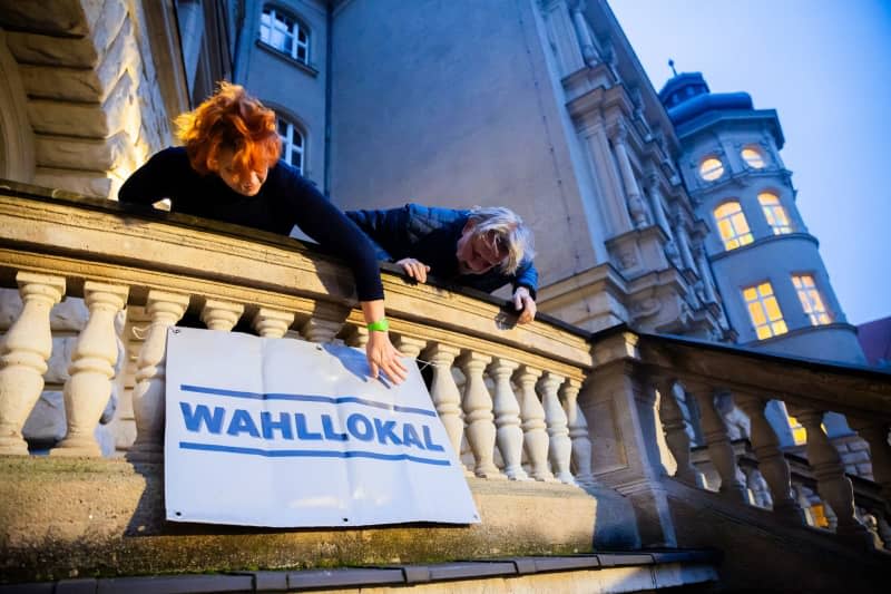 Two poll workers hang a sign at the polling station in the Carl-von-Ossietzky-Gymnasium in Berlin-Pankow before voting begins. Voters in hundreds of Berlin constituencies head to the polls for a partial re-run of the 2021 federal election, after numerous mishaps the first time round. Christoph Soeder/dpa