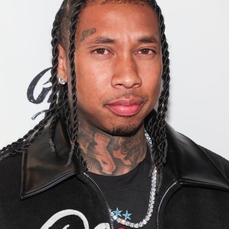 Tyga is seen at an event wearing a jacket with embroidery, a graphic T-shirt, a silver chain, and braided hair