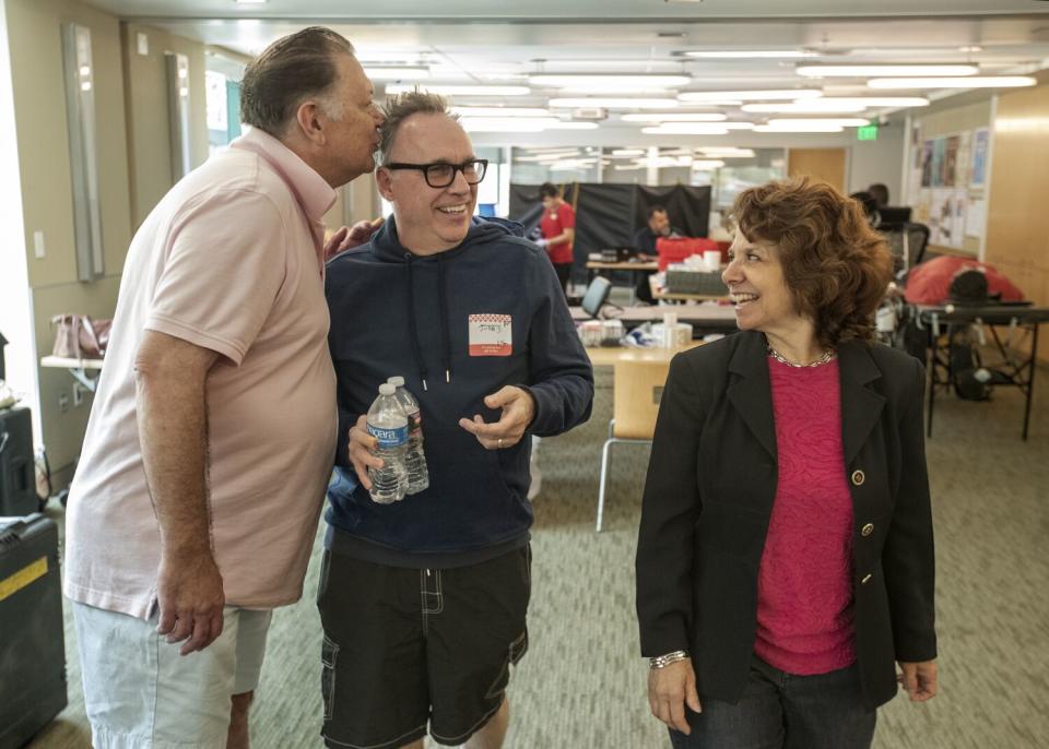 A man kisses another man on his head while a woman looks on, smiling.