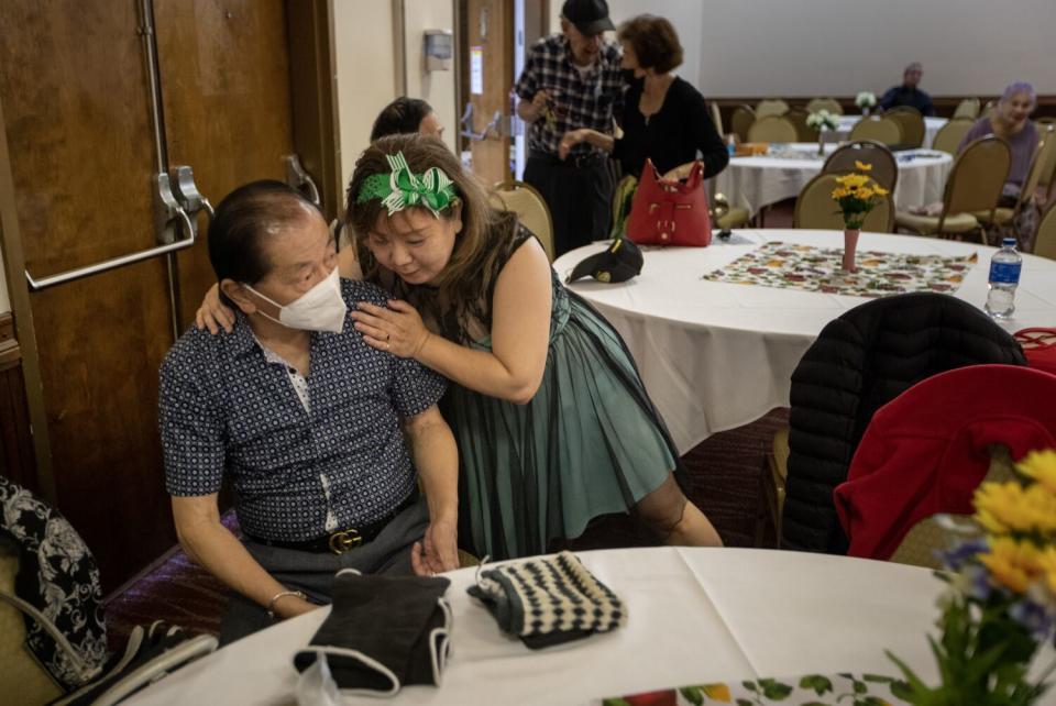 A woman leans over to give a side hug to a man who is seated and wearing a mask.