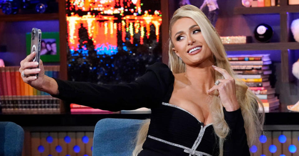 Paris Hilton sits on a talk show and takes a selfie with her phone. She is wearing a black and silver outfit.