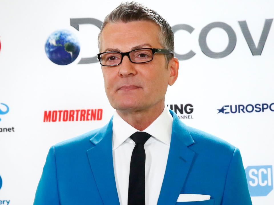 Randy Fenoli stands in a blue sit in front of a press background.