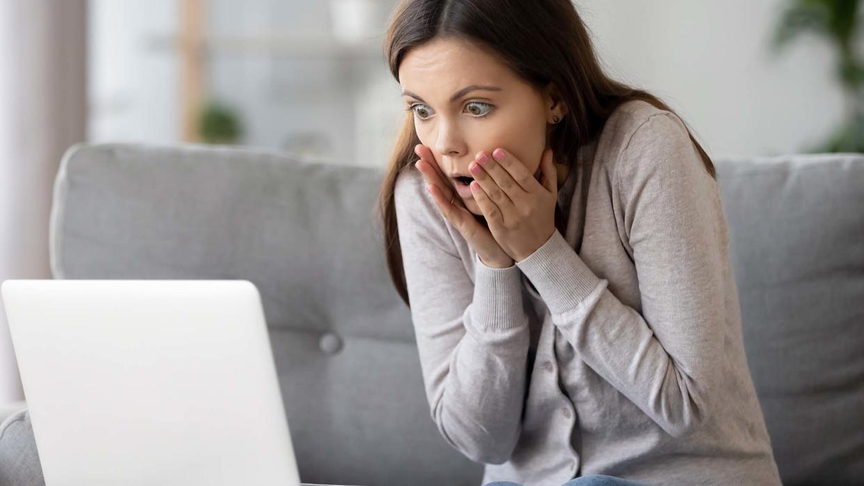  A shocked girl sitting on sofa at home looking on laptop screen. 