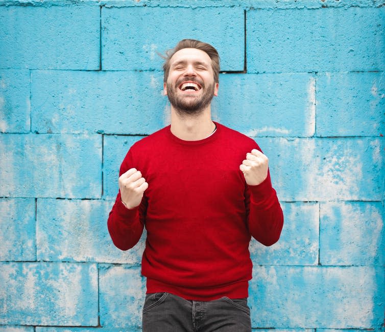 A grinning man in red jumper stands in front of blue wall.