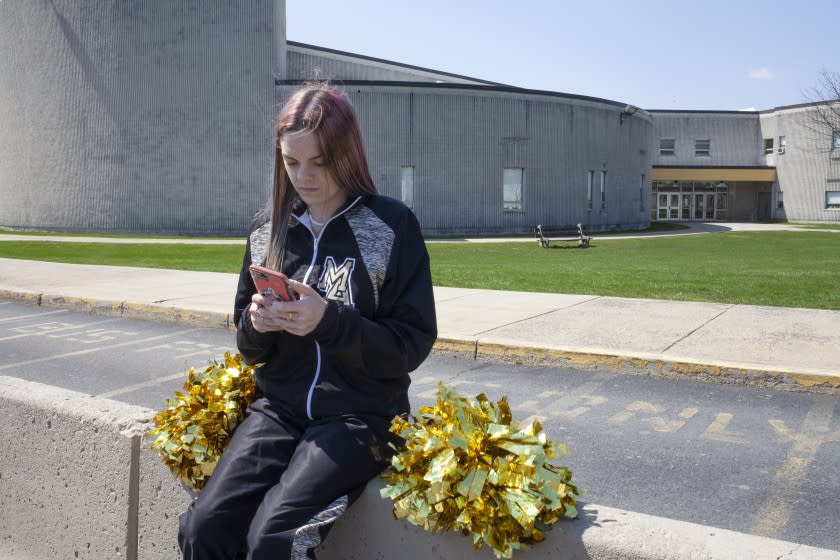 In this photo provided by the American Civil Liberties Union, Brandi Levy wears her former cheerleading outfit as she looks at her mobile phone while sitting outside Mahanoy Area High School in Mahanoy City, Pa., on April 4, 2021. A profanity-laced posting by Levy on Snapchat has ended up before the Supreme Court in the most significant case on student speech in more than 50 years. At issue in arguments to be heard Wednesday, April 28, 2021, via telephone, is whether public schools can discipline students over something they say off-campus. (Danna Singer/ACLU via AP)