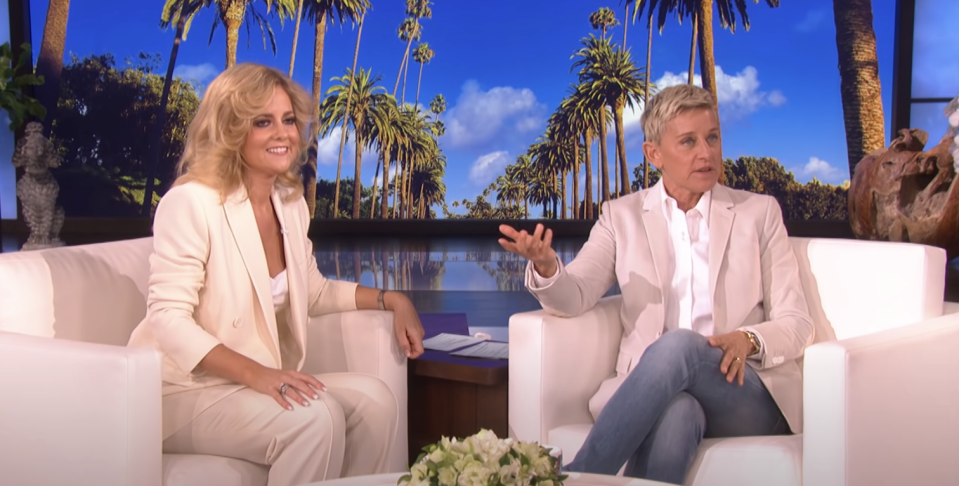 Two women, one in a white suit and another in a beige suit, having a conversation on a talk show set