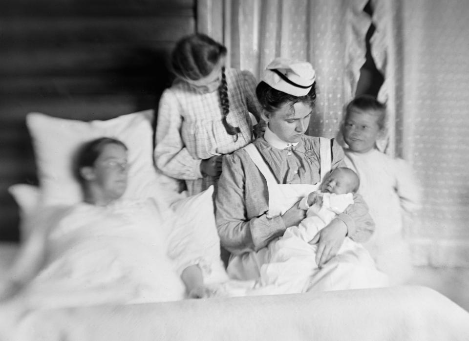 This vintage black-and-white photo shows a uniformed wet nurse holding a newborn baby surrounded by the birth mother, lying in bed, and the two new siblings.  