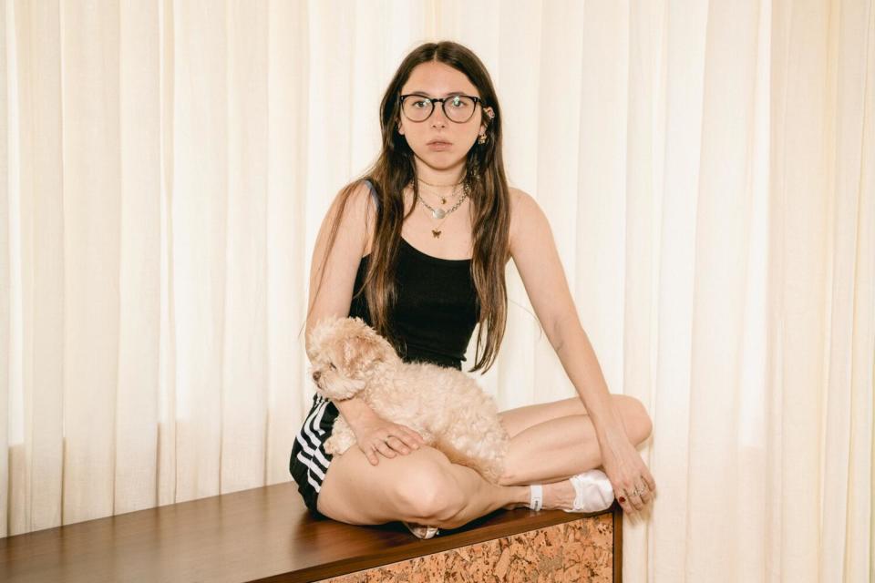 Esther Povitsky, wearing glasses, a tank top and athletic shorts, sits with her small, fluffy, cream-colored dog in her lap.