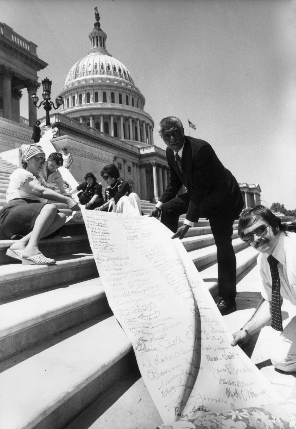 Democratic United States Representative Ralph Metcalfe of Illinois (C) holding a petition from his district measuring over 80-yards and with more than 10,000 names on calling for the impeachment of President Richard M. Nixon, Washington, DC. Metcalfe, along with eight other men and women are holding the petition on the steps of the Capitol Building.