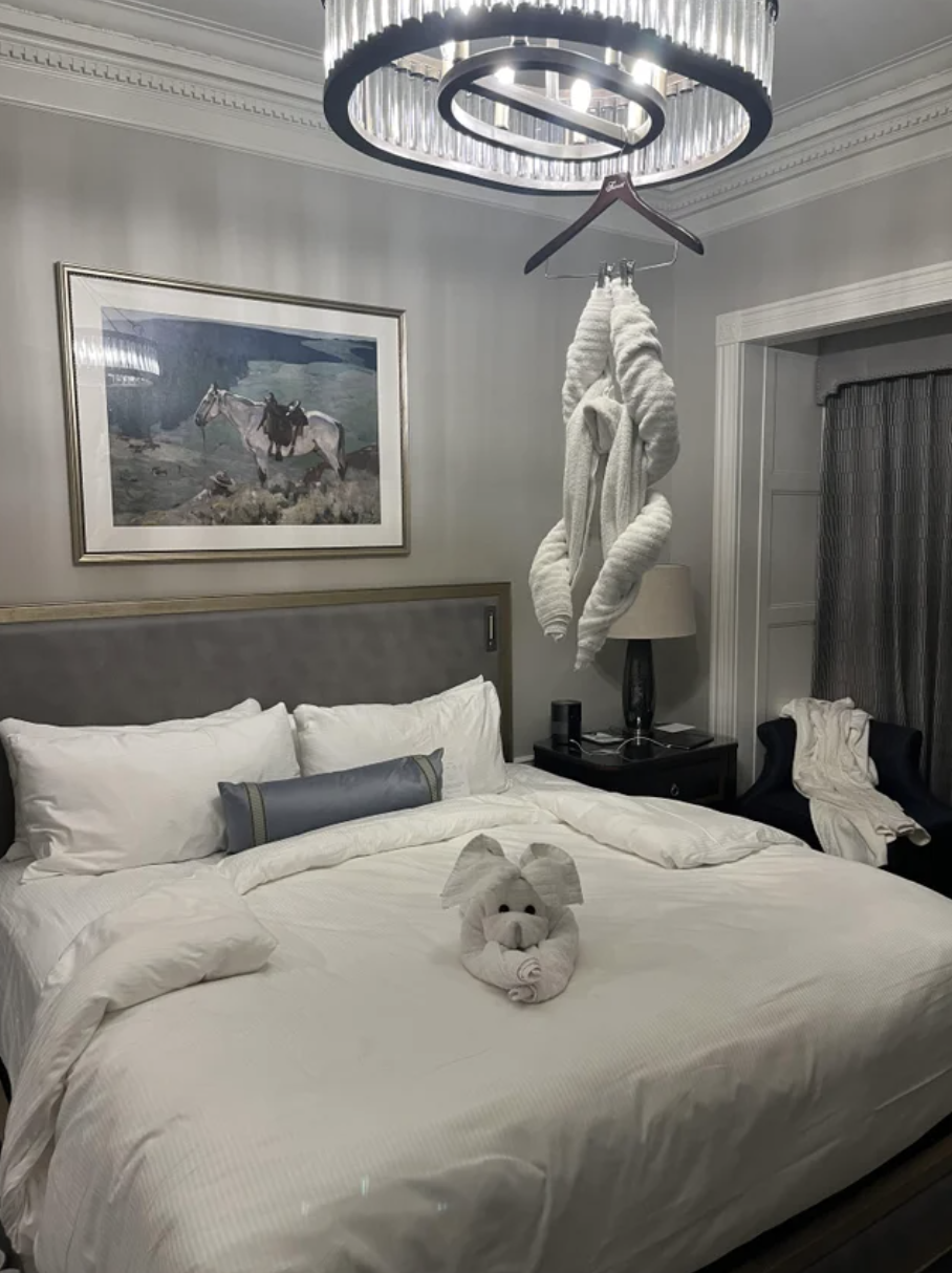 Towels loosely tied together in the shape of a person and hanging by the arms from a hanger attached to a ceiling light on the side of the bed, and more towels in the shape of an animal on the bed