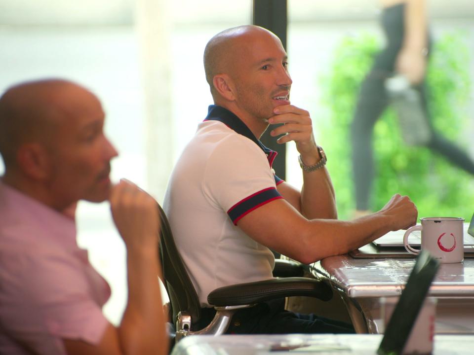 brett and jason oppenheim in an office, each sitting at desks with oppenheim group mugs and open laptops. they're posing similarly and holding their hands on their chins