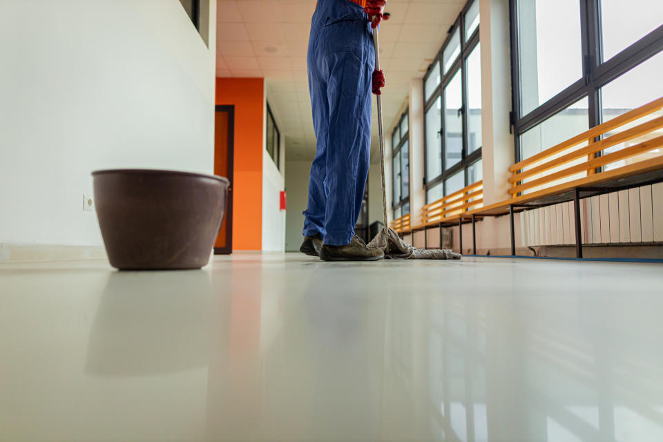 Person in blue coveralls mopping a glossy hallway floor, with a brown bucket nearby and benches along the windows