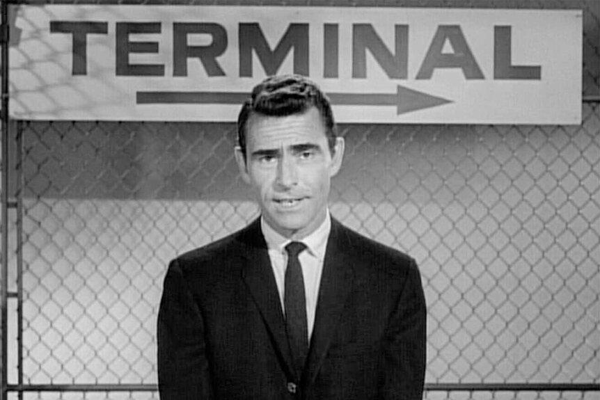 Rod Serling wears a suit and stands in front of sign that says 