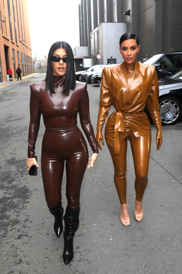 Kourtney in a chocolate brown latex catsuit with knee high black leather pointed boots. Kim in a similar caramel latex catsuit.
