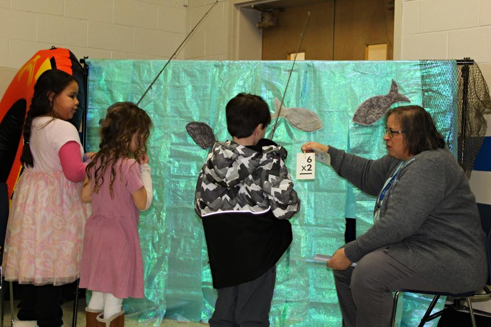 St. James Lutheran School first-grade teacher Lydia Roland holds up a multiplication problem for a child to solve during the STEM Fair Thursday, Jan. 18, 2024, in the school cafeteria. Kindergarten teacher Nikki Pollard hides behind the "water" and clips the math cards to each child's fishing pole.