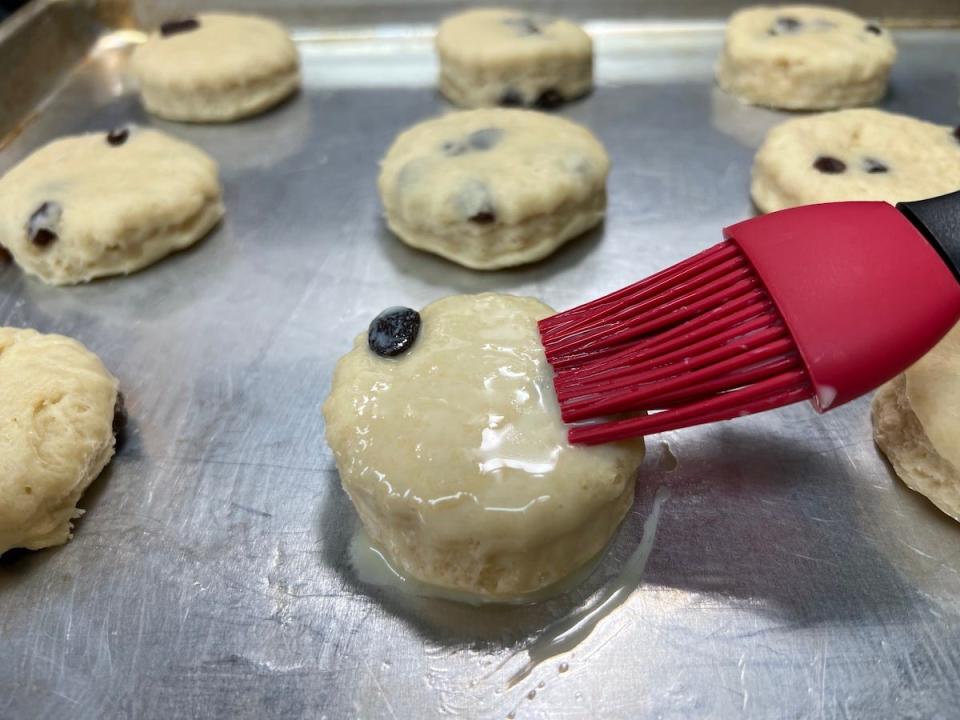 A close-up shot shows unbaked scones on a baking tray. A brush with egg wash is spread across one scone before they go in the oven.