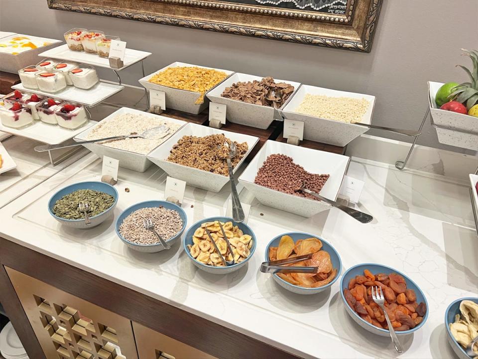 A white table with a breakfast buffet including grains, oatmeal, dried banana chips, dried apricots, and yogurt