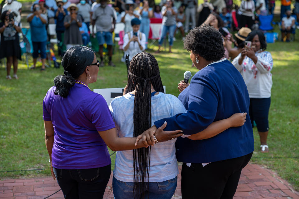 Stacey Abrams, with two other women, all of them with their arms around each other, speaks at a rally.