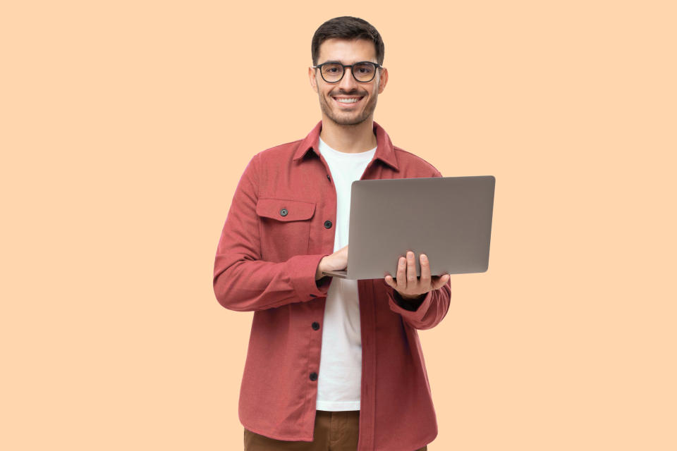 Smiling person holding a laptop standing against a plain background