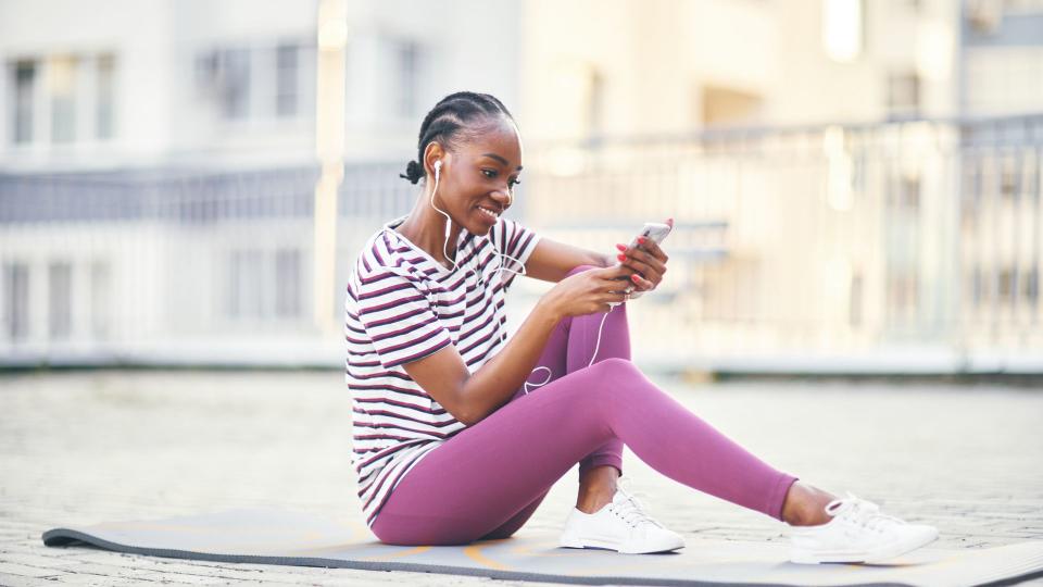 Smiling young woman in sports clothes on a yoga mat listening to music, side view