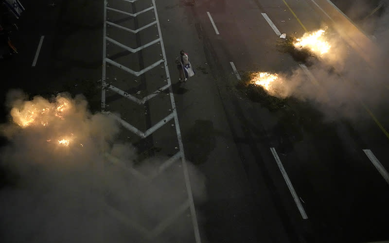 Bonfires burn as Israelis occupy the Ayalon Highway. The highway is seen from above and is dark and smoky, with three bonfires burning brightly and a single person standing nearby.