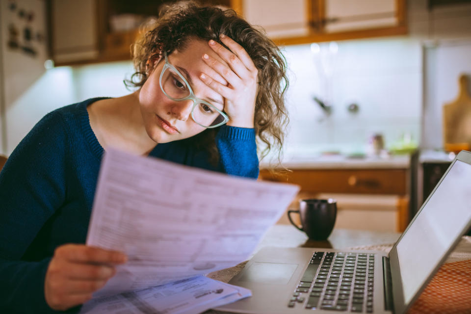 Person wearing glasses and a sweater looks stressed while reviewing financial documents next to a laptop in a kitchen