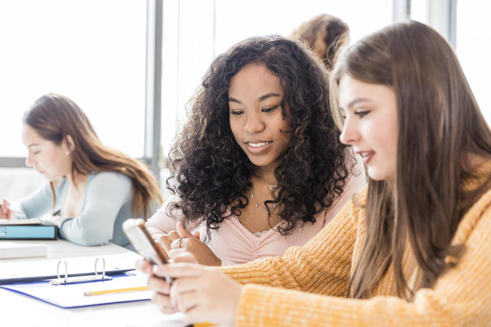 Three students at a table; two looking at a phone and one reading a book