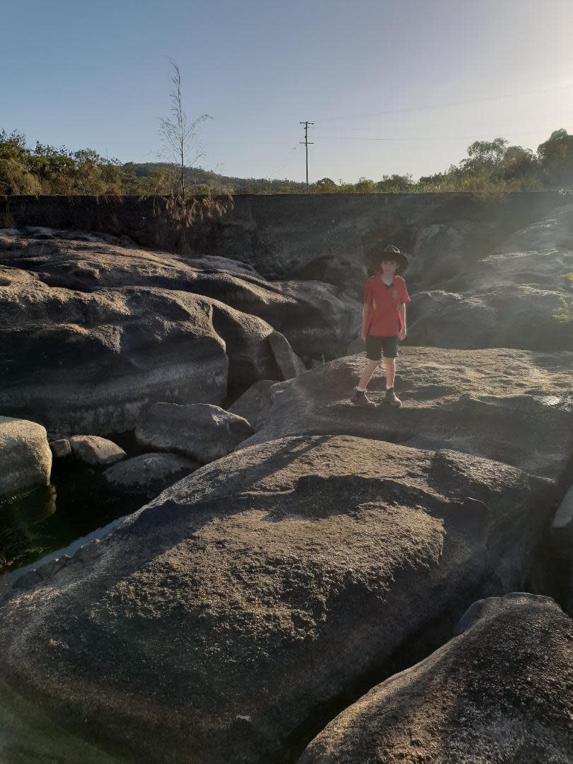 A young boy stands on a rock that forms part of a riverbed. 