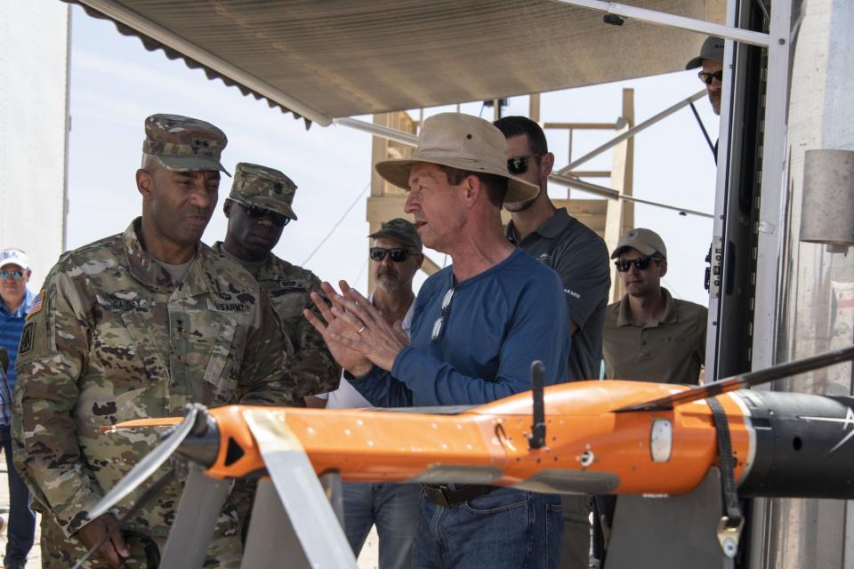 A man in civilian clothes talks to soldiers behind an orange drone.
