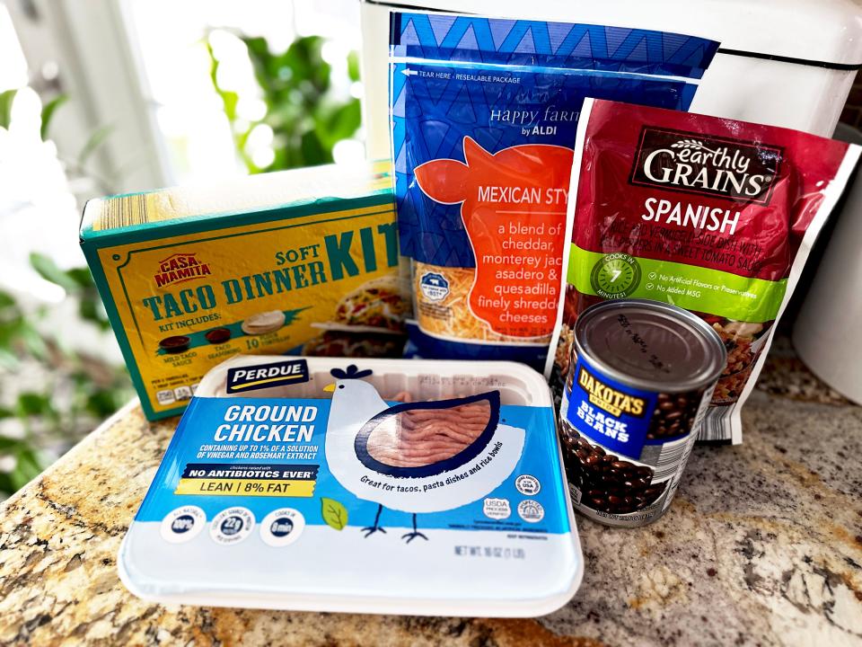 A container of ground chicken with blue labels, a can of black beans, a red pouch of Spanish rice mix, a blue and orange bag of shredded cheese, and a yellow and blue box with "taco dinner kit" text