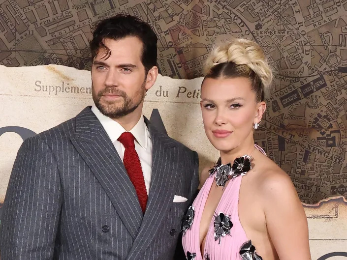 millie bobby brown and henry cavill on the enola holmes two red carpet. cavill is standing on the left, wearing a grey pinstripe suit with a red tie, and a coif of hair falling in his face. brown is wearing a pink halter gown with silver flowers on it, her blonde hair styled in a bun on top of her head.