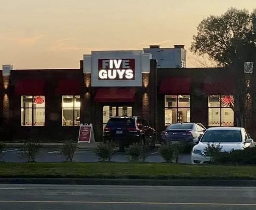 Five Guys restaurant exterior at dusk with a few cars parked outside. Twinkling lights are on inside the restaurant