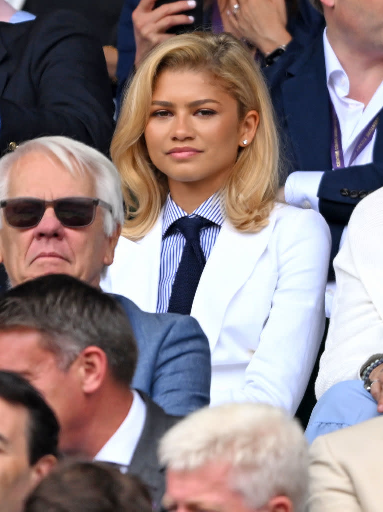 Zendaya seated among a crowd, wearing a jacket, striped shirt, and dark tie at an outdoor event with her hair down