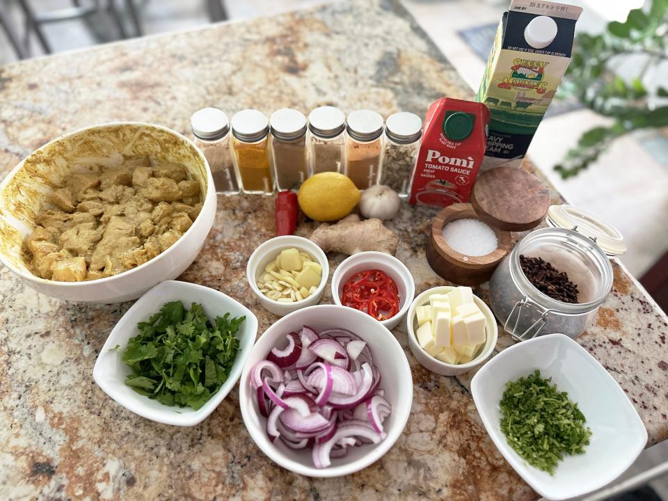 Several bowls of ingredients sit on a marble countertop. The ingredients include a bowl of raw chicken in a marinade, a bowl of cilantro, chopped red onion, a bowl of butter, bowls of chilis, a carton of tomato sauce, a carton of heavy whipping cream, and several spices lined up in a row