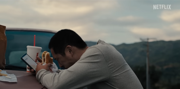 Steven Yeun as Danny leans over the hood of his car while eating Burger King in "Beef"