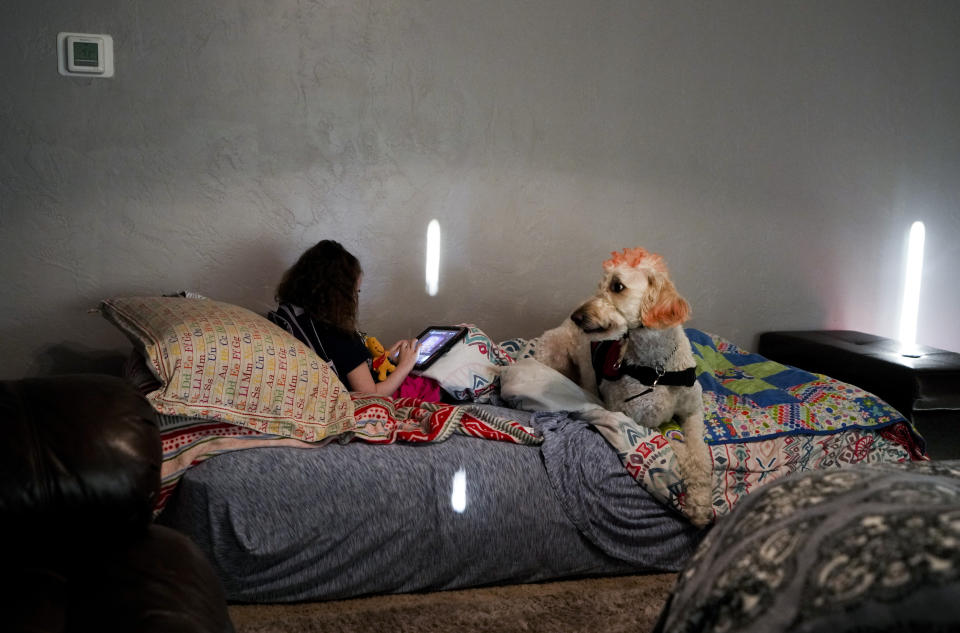 Scarlett Rasmussen, 8, sits at home with her tablet and service dog Riptide before getting ready for school, Wednesday, May 17, 2023, at their home in Grants Pass, Ore. Scarlett is nonverbal and uses an electronic device and online videos to communicate, but reads at her grade level. She was born with a genetic condition that causes her to have seizures and makes it hard for her to eat and digest food, requiring her to need a resident nurse at school. (AP Photo/Lindsey Wasson)