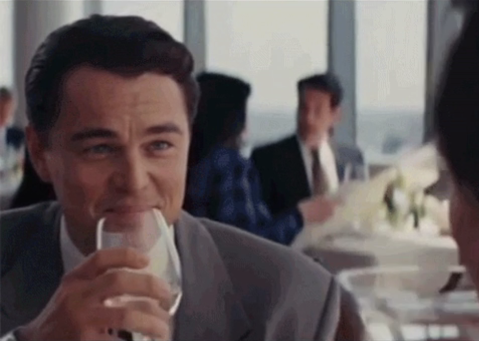 Leo DiCaprio in a suit smiling and raising a glass in a toast gesture in a social setting