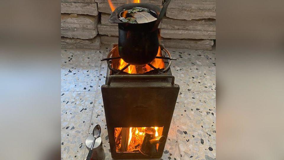 Rectangular black metal stove - with wood burning in bottom section and a pan on top. Placed on a tiled floor, with tongs and a spoon in front. 