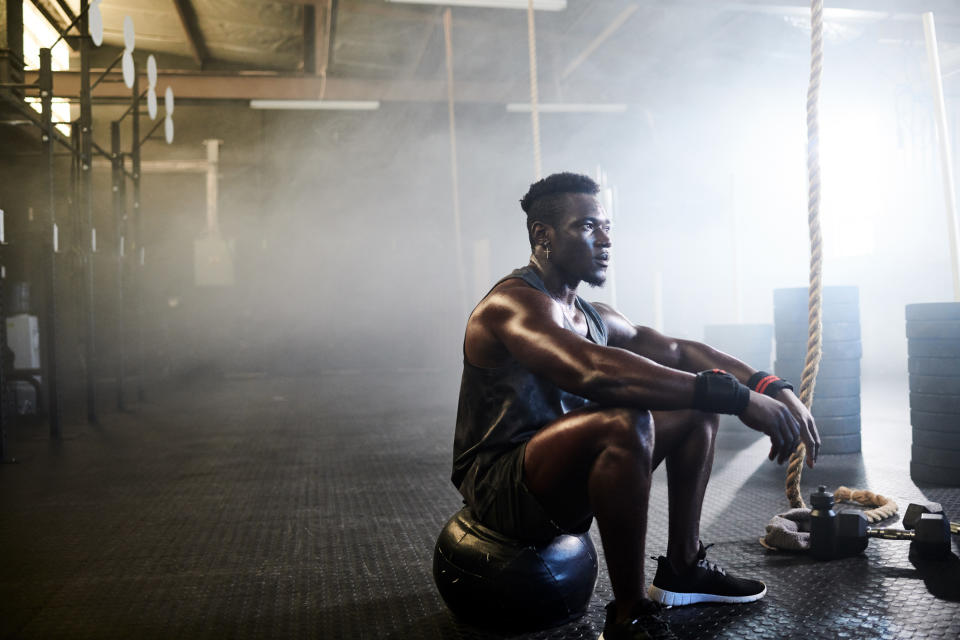 A person sits on a medicine ball in a gym, looking contemplative. They wear athletic clothing and sneakers, with gym equipment and foggy lighting in the background