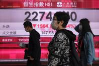 People wear protective masks as they walk past a panel displaying the Hang Seng Index during morning trading in Hong Kong