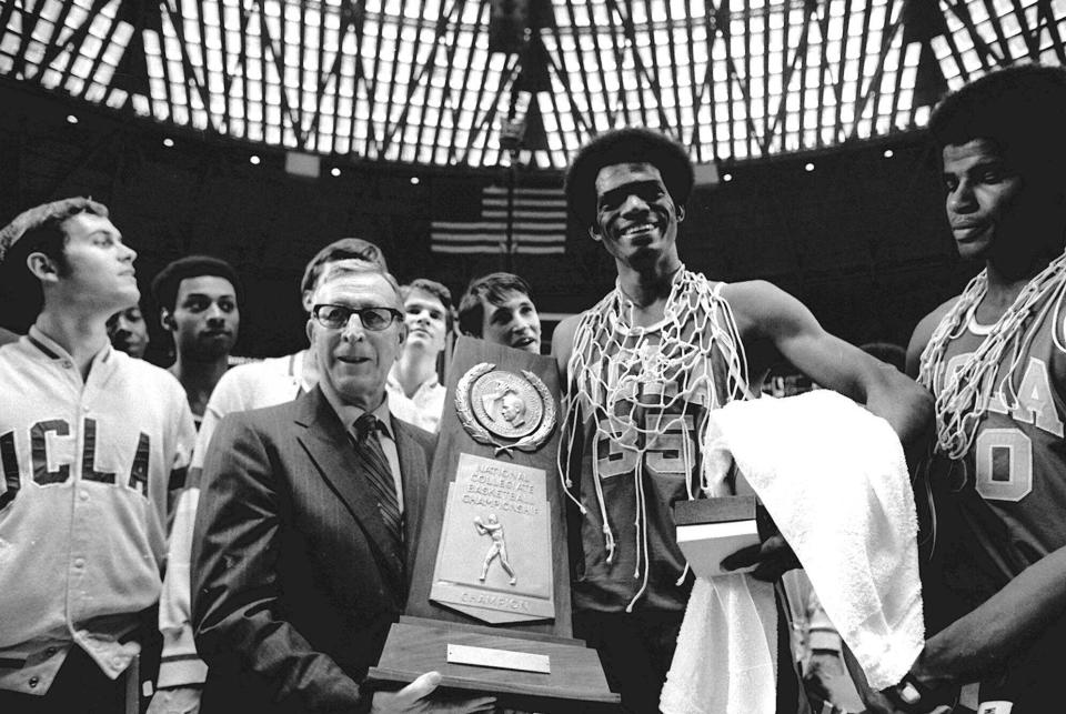 FILE - UCLA coach John Wooden holds the NCAA championship trophy which the Bruins won for the fifth straight year after beating Villanova 68-62 in Houston, March 27, 1971. Sidney Wicks (35) Bruins forward, wears part of the basket net around his neck. (AP Photo/File)