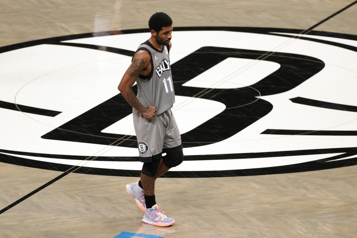 Kyrie Irving with his hands on his hips and looking down at the court near midcourt and the Brooklyn Nets logo.