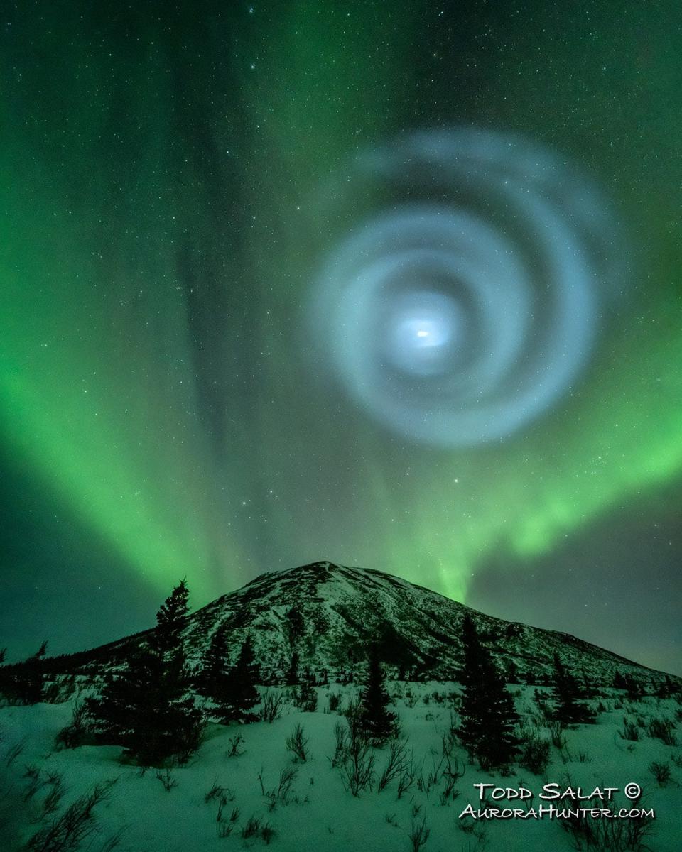 giant pale blue spiral again green stripes of the aurora borealis across a dark sky above a snowy mountain glowing green from the ligh