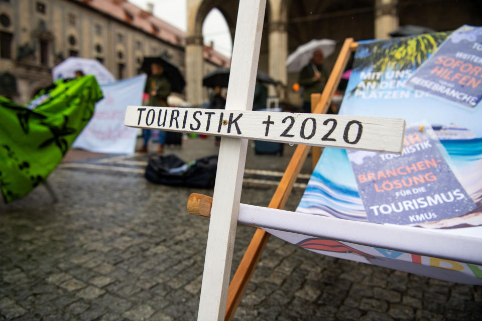 Bayern, München: "Touristik, gestorben 2020" steht auf einem Holzkreuz, das vor einer Sonnenliege im Regen steht. Reiseveranstalter haben bundesweit für einen Rettungsschirm gestreikt. Foto: Lino Mirgeler / dpa