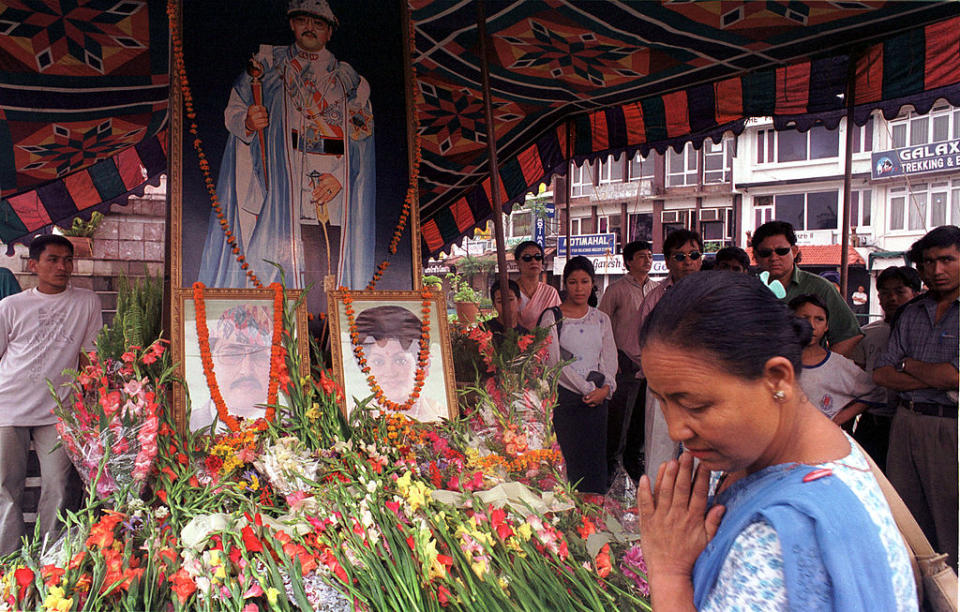 People gather at a shrine with flowers, featuring portraits and a large painting of King Birendra, paying respects in a solemn atmosphere