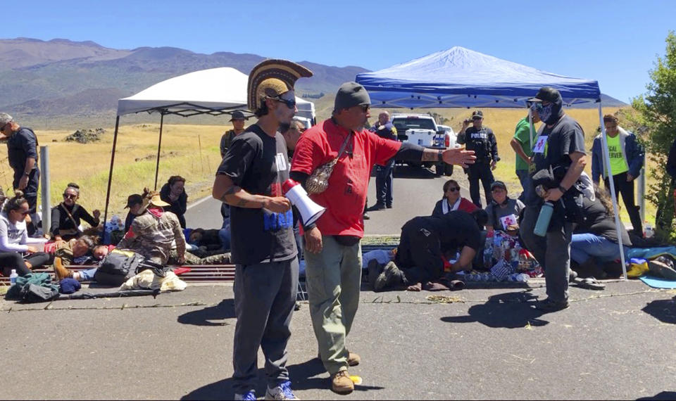 In this July 15, 2019, photo from video, protest leader Kaho'okahi Kanuha, center left, talks to demonstrators, some of whom have chained themselves to a cattle guard on a road at the base of Mauna Kea on Hawaii's Big Island. For activists who say they're protecting Mauna Kea, the fight against the proposed Thirty Meter Telescope is a boiling point in Hawaiian history: the overthrow on the Hawaiian kingdom, battles over land, water and development and questions about how the islands should be governed. (AP Photo/Caleb Jones)