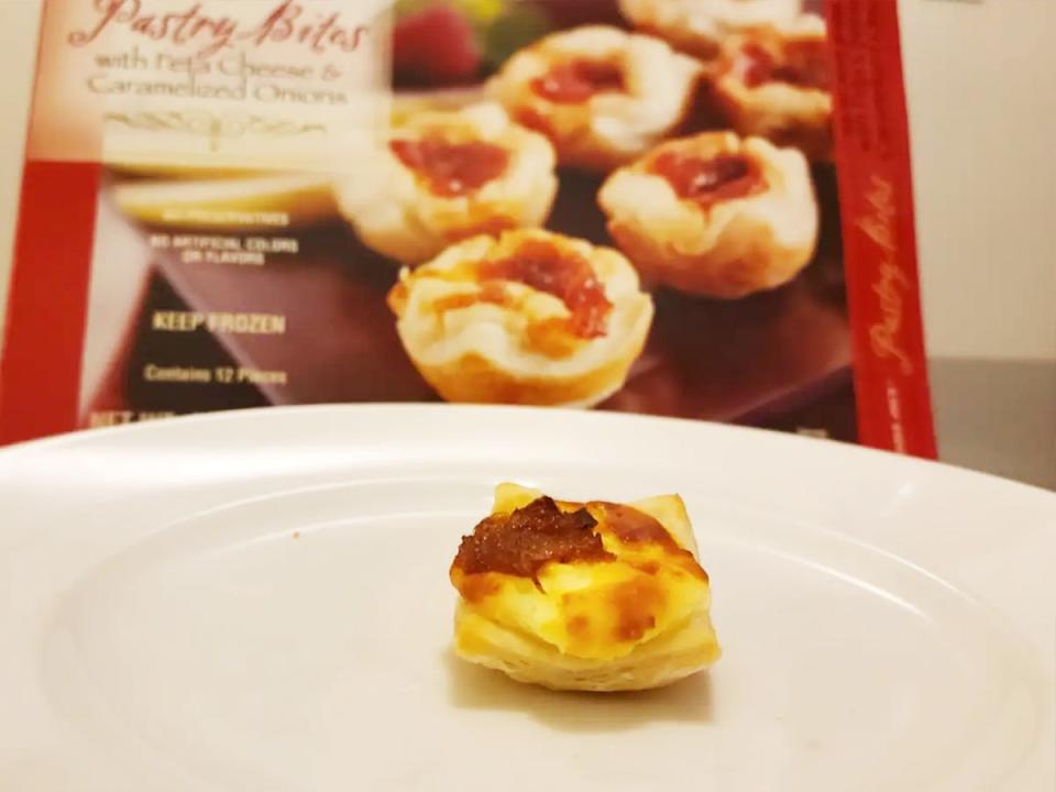 A golden-brown pastry bite with a darkened cheesy filling sits on a white plate with a red box behind the plate